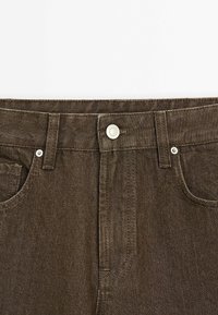 Bruine spijkerbroek taillesectie met metalen knoop, riemlussen en voorzakken, weergegeven op een witte achtergrond.