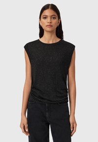 Donna con lunghi capelli scuri, che indossa un top senza maniche nero a macchie e pantaloni neri, in piedi contro uno sfondo grigio chiaro uniforme.