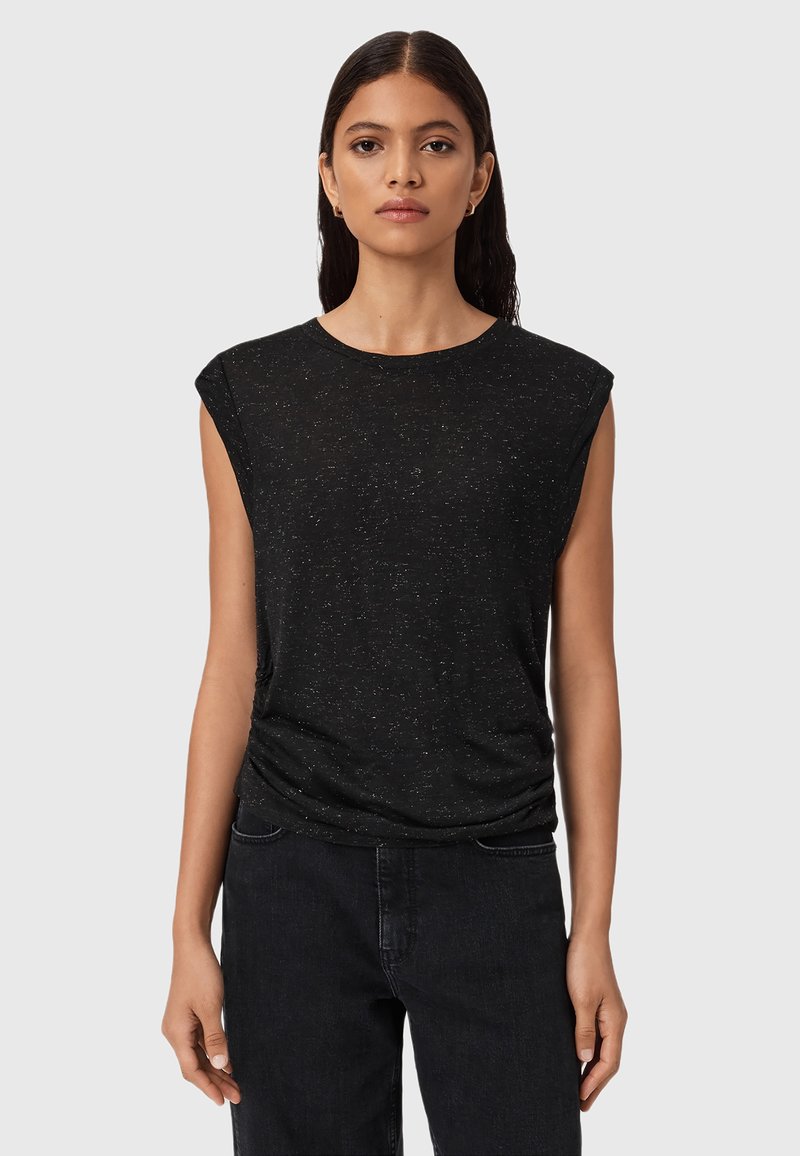 Donna con lunghi capelli scuri, che indossa un top senza maniche nero a macchie e pantaloni neri, in piedi contro uno sfondo grigio chiaro uniforme.
