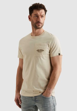 Moški, oblečen v bež kratek rokav majico z žepom spredaj in grafičnim napisom, stoji z eno roko v žepu svetlo modrih kavbojk.