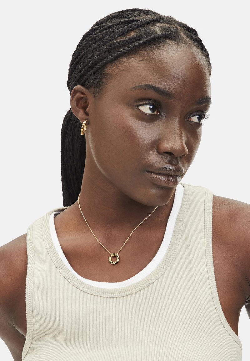 Goldschmuck mit einem kreisförmigen Anhänger im Sonnenstrahlendesign, kombiniert mit strukturierten Creolen, getragen über einem hellbeigen Tanktop.