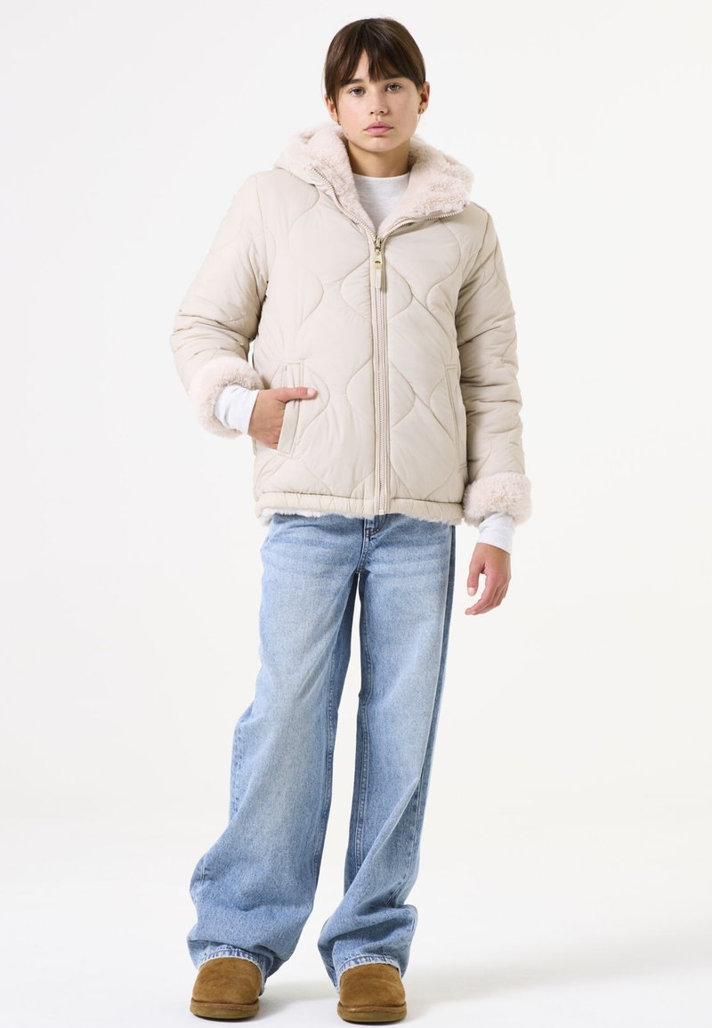 Chaqueta acolchada beige con capucha y puños de felpa, cremallera dorada y dos bolsillos frontales, combinada con jeans de pierna ancha de color azul claro y zapatos marrones.