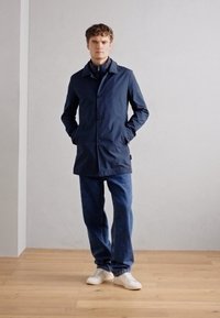 Uomo che indossa un cappotto blu navy con le mani in tasca, jeans blu e sneakers bianche, in piedi su un pavimento di legno davanti a una parete chiara e semplice.