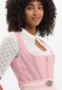 Rosa Cord-Minikleid mit Reißverschluss vorne, breiten Trägern und einer dekorativen silbernen Brosche in der Taille. Darunter eine weiße Spitzbluse.