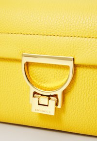 Borsa a tracolla gialla in pelle con superficie testurizzata, dotata di una chiusura in metallo color oro e il nome del brand "Coccinelle" impresso sulla chiusura.