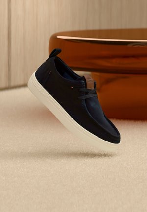 Marineblaue Freizeitsneaker mit weißer Sohle und braunem Lederakzent, schwebend über einem hellbeigen Teppich dargestellt.