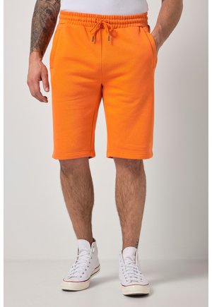 Mężczyzna ubrany w jasnopomarańczowe szorty ze sznurkiem, białe wysokie trampki oraz białą koszulkę, stojący na jednolitym tle.