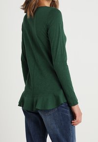Green long-sleeve top with a round neckline, featuring a flared ruffle hem and textured fabric. Paired with blue jeans.