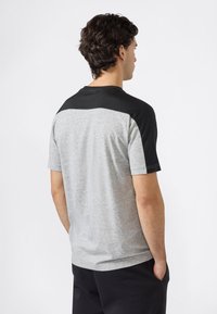 Joven con cabello rizado oscuro que lleva una camiseta de color gris y negro de manga corta y pantalones negros, de pie con las manos en los bolsillos.