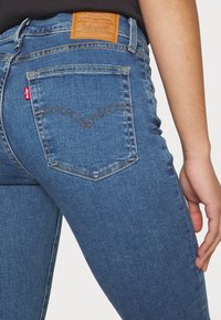 Zilās džinsu bikses ar piegriezumu, kas akcentē ķermeni, ar brūnu ādas etiķeti jostasvietā un sarkanu Levi's etiķeti aizmugurējā kabatā.