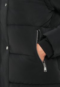 Fekete, steppelt pufferszövet kabát, oldalsó cipzáras zsebbel és rugalmas mandzsettákkal a tökéletes illeszkedésért. Simaságával és matt felületével tűnik ki.