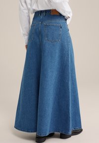 Modrá džínová maxi sukně s vysokým pasem, rozšířeným střihem a dvěma zadními kapsami. Lem je mírně roztrhnutý. Nosená s černými kotníkovými botami.