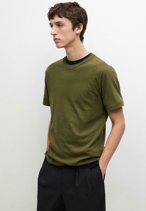 Giovane uomo con camicia verde oliva a maniche corte e pantaloni neri, in piedi con le mani nelle tasche contro uno sfondo chiaro e uniforme.