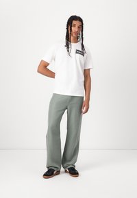 White t-shirt with black logo, paired with light green wide-leg pants and black shoes with brown soles, standing against a white background.