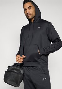 Μαύρη φούτερ από τη Nike με καγκουρό τσέπη και λευκό λογότυπο. Το μοντέλο κρατάει μία μαύρη τσάντα πλέγματος, φορώντας παντελόνι που ταιριάζει. Λείος, αθλητικός ύφασμα.