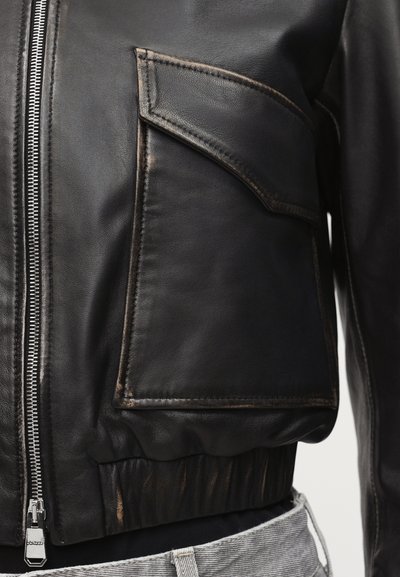 Close-up of black leather jacket pocket with flap and visible silver zipper, worn over light grey pants.