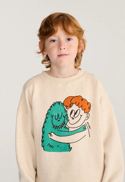 Enfant aux cheveux roux avec des taches de rousseur portant un sweat-shirt beige montrant un dessin animé d’un monstre vert aux cheveux orange qui fait un câlin.