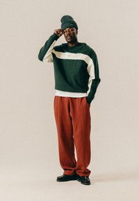 Πράσινη και κρεμ μπλοκαρισμένη φούτερ με ραμμένα μανίκια, σε συνδυασμό με χαλαρές κόκκινες παντελόνες και μαύρα παπούτσια, φορεμένα με μια ταιριαστή πράσινη σκούφια.