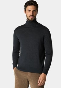 Uomo che indossa un dolcevita aderente grigio scuro e pantaloni in velluto a coste marrone chiaro, in piedi con una mano in tasca.