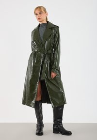Lesklý zelený trench coat s embosovaným vzorom krokodíla, s opaskom, širokými klopami a dĺžkou po kolená, v kombinácii s čiernymi čižmami po kolená.