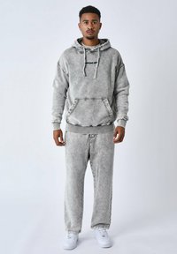 Sudadera gris desgastada con bolsillo canguro, combinada con pantalones de chándal grises a juego. Presenta costuras blancas y se complementa con zapatillas blancas.