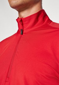 Veste zippée rouge en tissu lisse et léger. Dotée d'un col montant et d'une fermeture éclair avec une petite étiquette noire détaillant la marque.