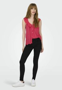 Blusa sin mangas rosa con cuello con volantes y cierre de botones, con detalles de ojetes; combinada con jeans ajustados negros y zapatillas blancas.