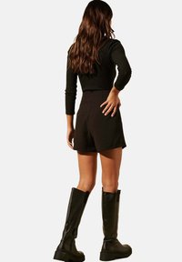 Haut noir à manches longues côtelé, short noir taille haute et bottes noires montantes. La mannequin a les cheveux ondulés et est vue de dos.