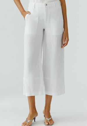 Femme portant un pantalon blanc coupe large et court avec poches plaquées à l'avant et des sandales argentées à talons, debout devant un fond uni.