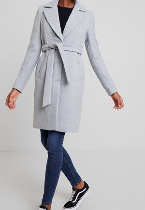 Woman wearing a light gray belted coat over dark jeans and black sneakers, standing with arms out and one leg crossed over the other.