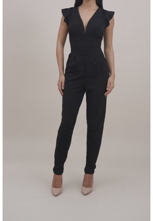 Kvinde iført en ærmeløs sort jumpsuit med flæse skuldre og dyb V-udskæring, kombineret med nøgenfarvede højhælede sko, står mod en ensfarvet baggrund.