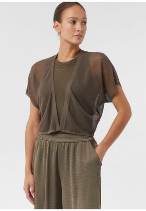 Kobieta w brązowym siatkowym kardiganie z krótkim rękawem, nałożonym na bezrękawnik, oraz w dopasowanych spodniach z wysokim stanem, stojąca z jedną ręką w kieszeni.