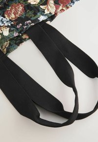 Svarta remmar av en blommönstrad tygväska ligger på en ljus bakgrund, vilket framhäver väskans intrikata mönster och textur.