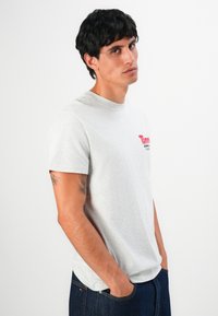 Jeune homme aux cheveux noirs courts portant un T-shirt gris clair de Tommy Jeans et un jean bleu foncé, les mains dans les poches, regardant légèrement sur le côté.