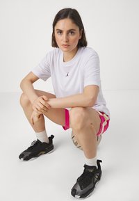 Hvit t-skjorte med svart logo, rosa shorts med gule striper, svarte treningssko, sittende på en fotball, nøytral bakgrunn.