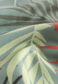 Tessuto decorato con foglie verdi e accenti rossi su uno sfondo testurizzato, che mostra un tessile morbido e drappeggiato con motivi naturali.