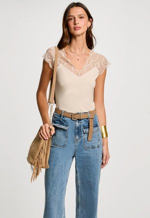 Beige top met kant mouwtjes, V-hals ontwerp, gecombineerd met lichtblauwe high-waisted jeans, geaccentueerd met een tan kleurige riem en een fringed tas.