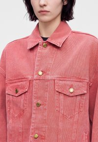 Personne portant une veste en denim rose délavée avec des boutons en laiton fermant le devant et les poches poitrine, debout devant un fond uni.
