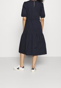 Marinblå bomullsklänning med puffärmar, insvängd midja och flerlagad kjol. Har en nyckelhålsdetalj på ryggen. Matchas med vita sneakers.