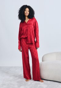 Set de pijamale din satin roșu, compus dintr-o cămașă cu nasturi cu model geometric și pantaloni largi. Textură netedă și croială lejeră.