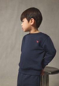 Granatowa bluza wykonana z miękkiego materiału, z okrągłym dekoltem i czerwonym logo na piersi, zestawiona z pasującymi spodniami.