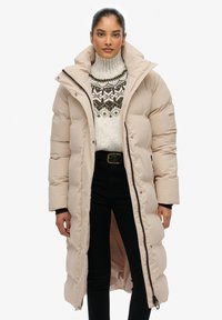 Beige pufferjack met een hoge kraag, ritssluiting en zijzakken, met een gestikt ontwerp en een middellange snit. Draagt over een patroontrui.