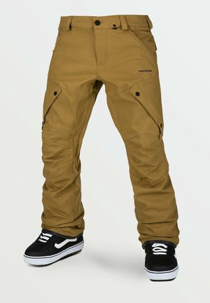 Pantalon cargo marron avec poches latérales boutonnées, genoux renforcés et baskets noir et blanc sur fond blanc.