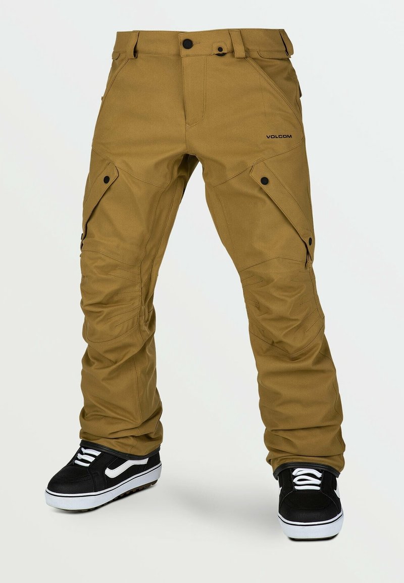 Pantalon cargo marron avec poches latérales boutonnées, genoux renforcés et baskets noir et blanc sur fond blanc.