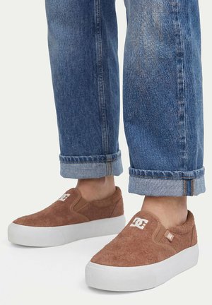Brune slip-on sneakers med hvide såler og DC-logo, båret med opkørte blå jeans.