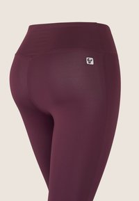 Leggings color burdeos con cintura alta, confeccionados en tela elástica. Presentan una pequeña etiqueta con logo en la parte trasera y una textura lisa y elegante.