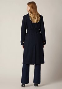 Granatowy płaszcz z paskiem o dopasowanym kroju, zdobiony złotymi guzikami, o długiej sylwetce i gładkim wełnianym wykończeniu. Widać szerokie jeansy i botki na obcasie.