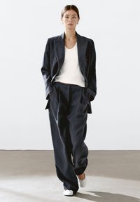 Completo scuro su misura con giacca doppiopetto, pantaloni a gamba larga e top bianco. Tessuto liscio, design minimale e sfondo neutro.