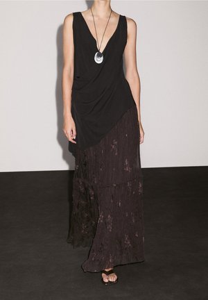 Femme portant un haut noir sans manches à nouer, une longue jupe texturée foncée, des sandales noires, et un collier pendentif circulaire en argent, debout à l'intérieur.