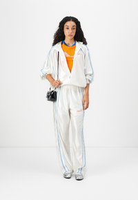 Giovane donna che indossa una tuta bianca con cerniera e piping blu, una maglietta arancione, scarpe argentate e porta una piccola borsa nera, in piedi su uno sfondo bianco.